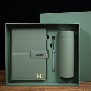 Puede incluir: Un juego de regalo verde salvia que contiene un cuaderno, un bolígrafo y un vaso térmico. El cuaderno tiene un cierre de solapa y las iniciales "MJ" en dorado. El bolígrafo y el vaso son del mismo color que el cuaderno. El juego se presenta en una caja verde a juego.