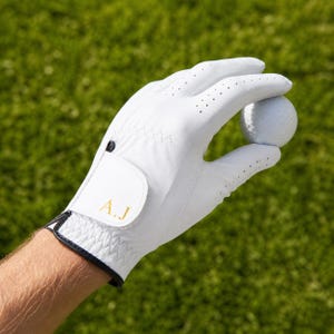 Può includere: Guanto da golf bianco con finiture nere e iniziali dorate "A.J". Il guanto tiene una pallina da golf bianca. Lo sfondo è erba verde.