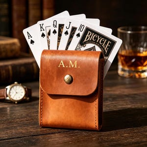 May include: A tan leather card holder, embossed with the initials "A.M." in gold, containing playing cards. The cards are fanned out, showing the Ace, King, Queen, Jack, and 10 of Spades. A watch and a glass of amber liquid are in the background.