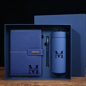 May include: A blue gift set containing a notebook, pen, and insulated tumbler. The notebook features a flap closure and white stitching. The tumbler and notebook are printed with the name "MICHAEL" and the initials "MVI". The set is presented in a matching blue box.