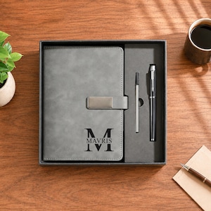 May include: A grey notebook with a magnetic closure and the name MAVRIS printed in black, presented in a black gift box alongside a black pen and a silver ink refill. The notebook cover features a large letter M above the name MAVRIS.
