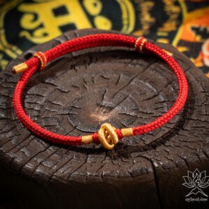 May include: A red woven bracelet with gold-colored accents. The bracelet is displayed on a dark brown, textured surface. The bracelet features a central gold-colored clasp and gold-colored details at the ends.