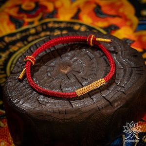 May include: A red bracelet with gold accents. The bracelet is made of a woven red cord with gold-colored metal details at the ends and in the center. The bracelet is displayed on a dark wooden surface.