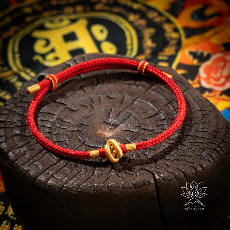 May include: A red woven bracelet with gold-colored accents. The bracelet is displayed on a dark brown, textured surface. The bracelet features a central gold-colored clasp and gold-colored details at the ends.