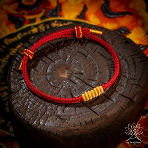 May include: A red and gold woven bracelet. The bracelet is made of a red cord with gold accents. The bracelet is displayed on a dark brown, textured wooden surface. The bracelet is a simple, elegant design.