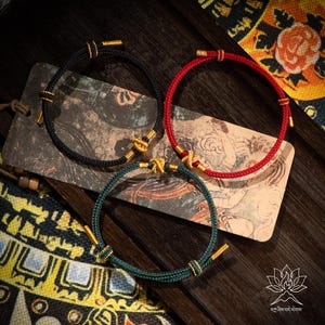 May include: Three braided cord bracelets in black, red, and green, each with gold-coloured accents. The bracelets are displayed on a decorative card with an Asian-inspired design, set against a dark wooden surface. The bracelets are a stylish accessory.