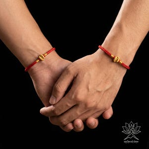 May include: Two red string bracelets with gold-coloured clasps are visible on wrists. The bracelets are worn on both wrists. The background is black.