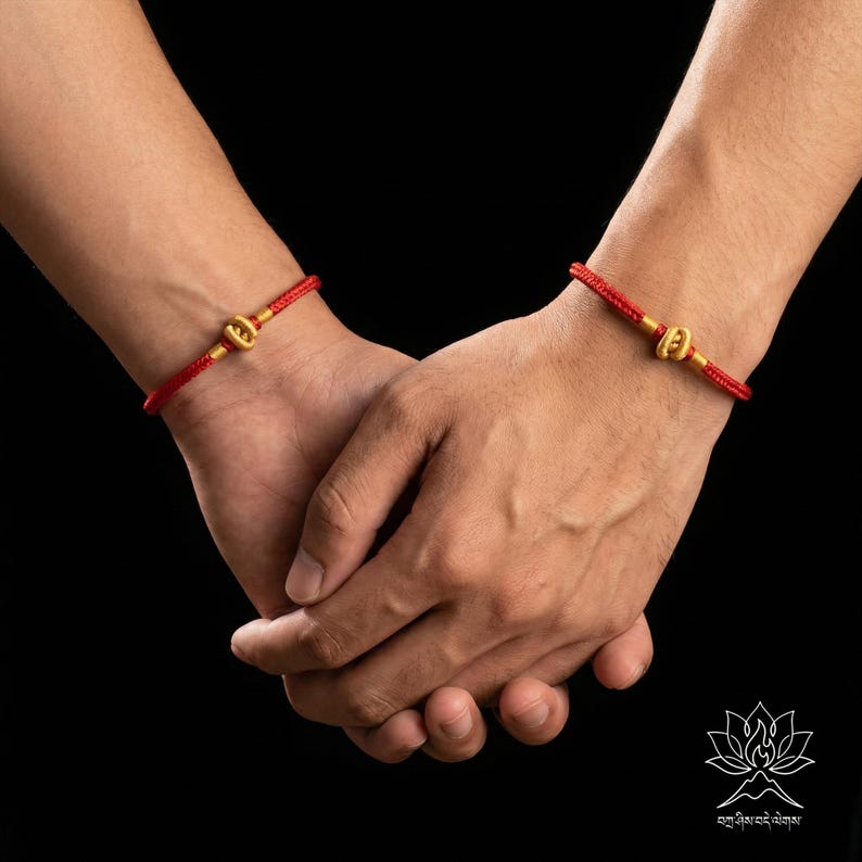 May include: Two red string bracelets with gold-coloured clasps are visible on wrists. The bracelets are worn on both wrists. The background is black.