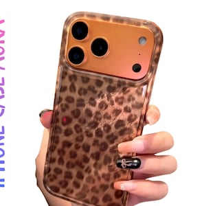May include: A clear iPhone case with a leopard print design. The case has a brown and tan pattern and a clear border. The phone's camera is visible through the case. The text "IPHONE CASE AURA" is printed on the left side of the case.