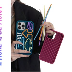 Könnte beinhalten: Türkisfarbene iPhone-Hülle mit farbenfrohem, abstraktem Design und eine magenta Hülle mit gepunkteter Textur. Der Text "IPHONE CASE AURA" befindet sich auf der linken Seite des Bildes. Die Hüllen werden mit bunten Stäben gehalten.