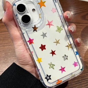 Può includere: Custodia trasparente per telefono con interno bianco e dettagli a forma di stella colorati. La custodia ha un bordo trasparente e un ritaglio per la fotocamera. Le stelle sono di vari colori, tra cui oro, rosa e rosso. La custodia è progettata per proteggere uno smartphone.