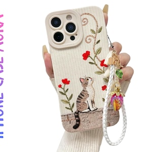 Peut inclure: Coque de téléphone beige avec broderie de chat, fleurs rouges et feuilles vertes. La coque a une dragonne tressée blanche avec un charme. Le texte "IPHONE CASE AURA" est sur le côté gauche.