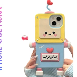 Può includere: Custodia per iPhone a forma di robot, nei colori giallo e blu, con dettagli rosa e un'antenna a forma di cuore. Il robot ha una faccia sorridente e un cuore sul petto. Il testo "iPHONE CASE AURA" è visibile.