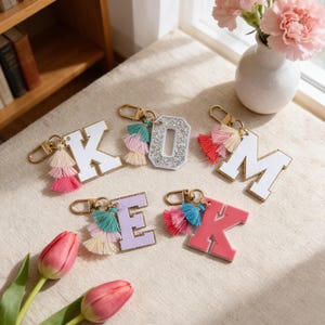 May include: A collection of letter and number keychains with colorful tassels. The letters K, O, M, E, and K are displayed in various colors, including white, pink, and lavender. The letter O is embellished with rhinestones. Each keychain has a gold-colored clasp.