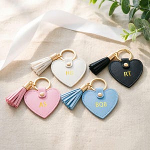May include: Heart-shaped keychains in white, black, pink, and blue with gold key rings and tassels. Each keychain has initials in gold lettering. The keychains are arranged on a neutral surface.
