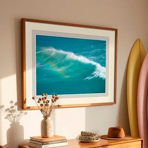 Puede incluir: Una impresión de arte mural horizontal enmarcada que representa una ola oceánica turquesa con un arcoíris, sobre una cómoda de madera. La cómoda sostiene un jarrón moteado con flores secas, libros, un cuenco pequeño y un sombrero fedora marrón.