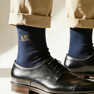 Può includere: Calzini blu navy con iniziali "AR" ricamate in oro sono indossati con scarpe eleganti in pelle nera. Le scarpe hanno un design a punta e sono abbinate a pantaloni color cachi. Primo piano sulla trama della calza.