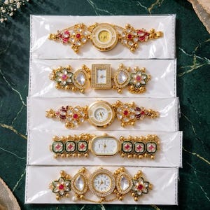 Puede incluir: Cinco relojes de pulsera dorados con detalles decorativos inspirados en flores, con piedras rojas y transparentes incrustadas. Cada reloj está empaquetado individualmente en una funda de plástico transparente, mostrando metalurgia y formas variadas.