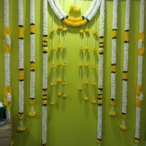 Op de afbeelding: Een decoratieve bloemenslinger met witte, gele en zwarte accenten. Het ontwerp omvat lange, verticale strengen en een gebogen bovendeel, alles tegen een groene achtergrond. De slinger wordt waarschijnlijk gebruikt voor feestelijke gelegenheden.