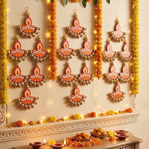 May include: A wall decoration featuring multiple hanging ornaments shaped like diya lamps with orange and red patterns, accented by gold-toned beads. The display is framed by yellow marigold garlands and string lights, with a wooden table holding bowls of sweets.