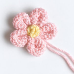 May include: A five-petaled crochet flower applique made from soft pink yarn with a bright yellow center. The flower features a textured, looped stitch pattern and includes two long, loose pink yarn strands extending from the bottom edge.