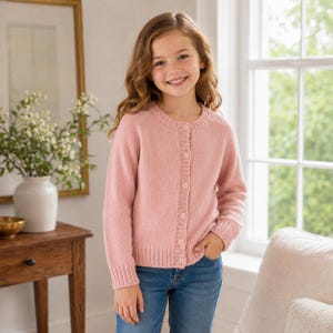 May include: A long-sleeved, light pink knit cardigan featuring a round neckline, a vertical button-down front with matching pink buttons, and ribbed cuffs and hem. The garment is worn over blue denim jeans, showcasing a simple, classic design.