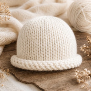 May include: A cream-coloured knit beanie with a rounded crown and a thick, textured rolled brim. The hat is constructed from a soft, chunky yarn featuring a vertical stockinette stitch pattern throughout the main body and a braided edge.