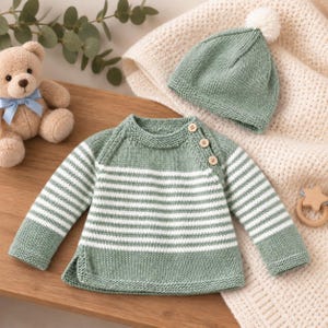 Puede incluir: Un suéter de punto a rayas verde salvia y blanco con tres botones de madera en el hombro y un gorro de punto verde salvia a juego con un pompón blanco. El suéter presenta mangas largas y un cuello acanalado, junto a un gorro de punto suave.