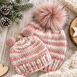 Puede incluir: Dos gorros de punto a juego con un patrón de rayas en tonos rosa, blanco y beige. El gorro más grande tiene un pompón grande y esponjoso de piel sintética rosa, mientras que el más pequeño presenta dos pompones de lana redondos y pequeños.