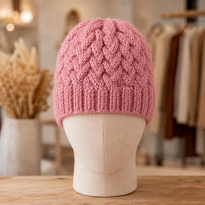May include: A dusty pink knit beanie featuring a prominent braided cable pattern across the crown and a ribbed cuff at the base. The hat is displayed on a neutral-toned mannequin head, showcasing the textured yarn construction and soft colour.