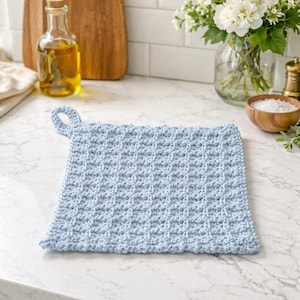 May include: A light blue, square crochet potholder with a loop for hanging. The potholder has a textured pattern and is displayed on a white countertop. Kitchen and home decor.