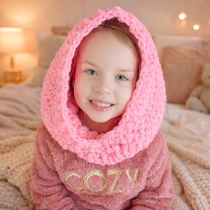 Könnte beinhalten: Eine junge Person trägt einen rosa, grob gestrickten Rundschal. Die Person trägt auch ein rosa, flauschiges Langarm-Oberteil mit dem Wort "COZY" in Goldfaden gestickt. Der Hintergrund umfasst ein Bett und sanfte Beleuchtung.