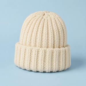 May include: A cream-colored beanie hat featuring a thick, vertical ribbed knit pattern. The hat has a wide, folded cuff at the base, creating a structured, rounded silhouette. The texture is consistent throughout, showcasing a chunky yarn construction.