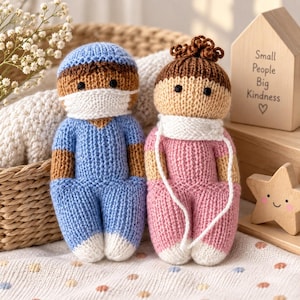 May include: Two handmade knitted dolls. One doll wears a blue scrub top and pants with a white face mask. The other doll has brown hair in a bun, a white scarf, and a pink outfit. A wooden house-shaped sign reads Small People Big Kindness.