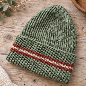 Könnte beinhalten: Eine salbeigrüne Strickmütze mit einem breiten, umgeschlagenen Bündchen. Das Bündchen ist mit einem horizontalen Streifenmuster verziert, das aus einer dünnen cremefarbenen Linie zwischen zwei dunkelroten Linien in grober Rippstrickoptik besteht.