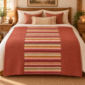 Op de afbeelding: Een gehaakte bedloper met een breed middenpaneel van horizontale strepen in tinten terracotta, crème, roze en saliegroen, geflankeerd door effen terracotta-kleurige gehaakte zijpanelen, gedrapeerd over een witte sprei.