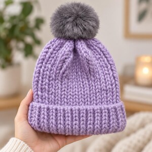 May include: A chunky knit beanie in a light purple color featuring a thick ribbed cuff and a large, fluffy dark gray faux fur pom-pom attached to the top. The hat is constructed with a vertical stitch pattern and a folded brim design.