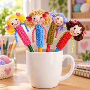 May include: Four pens topped with crocheted doll figures in a white ceramic mug. The dolls feature yarn hair in yellow, blonde, purple, and brown, with matching crocheted outfits in pink, blue, green, and red, each with embroidered facial features.