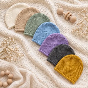 May include: A collection of eight knitted beanies in various solid colors including cream, tan, sage green, light blue, lavender, dark gray, and mustard yellow, arranged in a diagonal row on a textured white fabric surface with a wooden rattle.