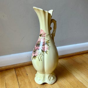May include: A pale yellow ceramic vase featuring a bulbous base, a slender neck with a flared rim, and a decorative handle. The front of the vase is adorned with a floral illustration of pink anemone flowers with green stems and leaves.