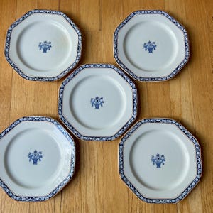 May include: A set of five off-white octagonal ceramic plates, each featuring a central blue floral basket motif and a decorative blue and white patterned border along the rim. The plates are arranged in a cluster on a wooden surface.