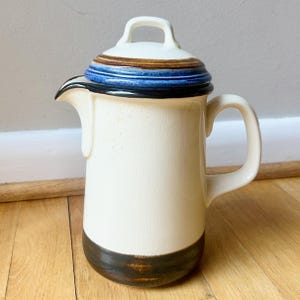May include: A ceramic coffee pot with a cream-colored body, featuring a dark brown base and a lid decorated with bands of brown and blue glaze. The pot has a curved handle, a prominent spout, and a small handle on top of the lid for easy removal.