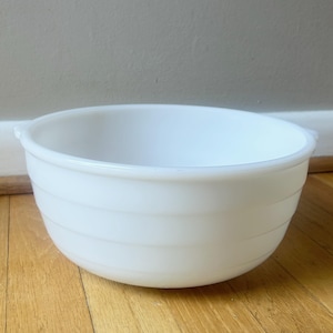 May include: A white, round mixing bowl with horizontal ridges. The bowl has a smooth, glossy finish and small handles on either side. It sits on a wooden surface.