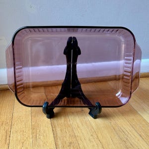 May include: A rectangular purple glass baking dish with integrated side handles and a ribbed exterior edge design. The center of the dish features embossed text that reads Pyrex, Made in USA, and includes specific model and capacity markings.