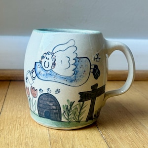 May include: Hand-painted ceramic mug with a whimsical design. The mug features a cartoon angel in a blue dress, a beehive, flowers, and a signpost. The mug has a light blue rim and a curved handle.