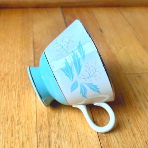 May include: A light blue teacup featuring a white band decorated with a pattern of light blue leaves and small pink berries. The teacup has a white handle and a gold-toned rim along the top edge, resting on its side on a wooden surface.