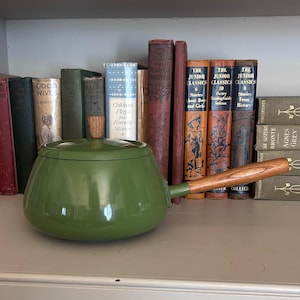 May include: A green enamel saucepan with a wooden handle and lid, set against a backdrop of vintage books. The saucepan has a rounded shape and a long handle. The books are of various sizes and colors.