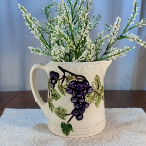 May include: Cream-colored ceramic pitcher with a handle, featuring a raised design of purple grapes and green leaves. The pitcher is filled with white floral stems. The pitcher has a decorative geometric pattern around the top and bottom.
