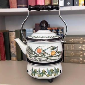 May include: White enamel tea kettle with a black rim and handle. The kettle features a botanical design with oranges and the text "Le Bunch". The kettle sits on a matching warmer with a similar design. A classic kitchenware item.