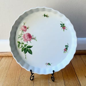 May include: White ceramic pie dish with a scalloped edge, adorned with pink rose illustrations and green leaves. The dish is displayed on a black metal stand, showcasing a classic floral design.